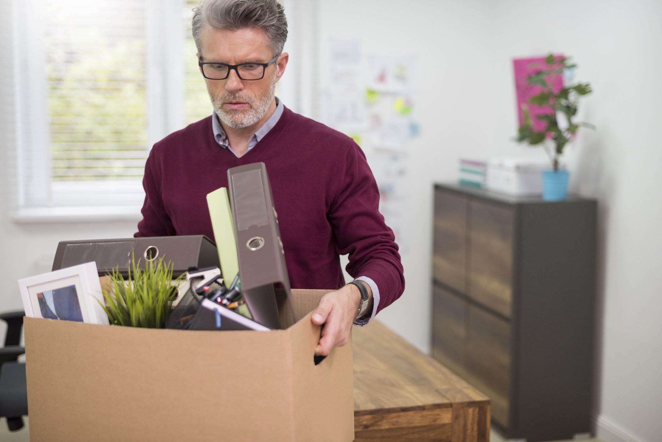 a sad man carries a box of his belongings out of an office