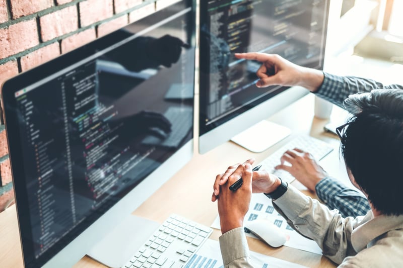 employees hands point at dual computer screens with code on them