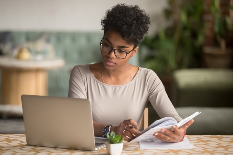 a woman with short dark hair holds a notebook and pen and looks to her right to her laptop screen