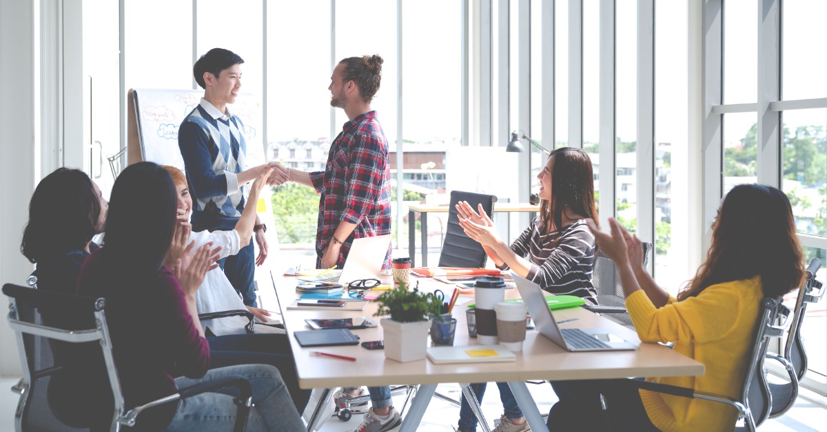 Employees sitting around table with two people standing and shaking hands