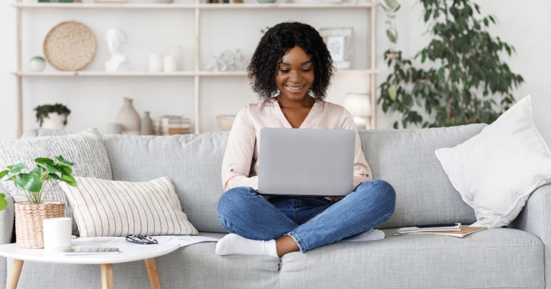 Remote Work. Millennial african american woman working on laptop at home, sitting on couch in living room with stylish interior, free space