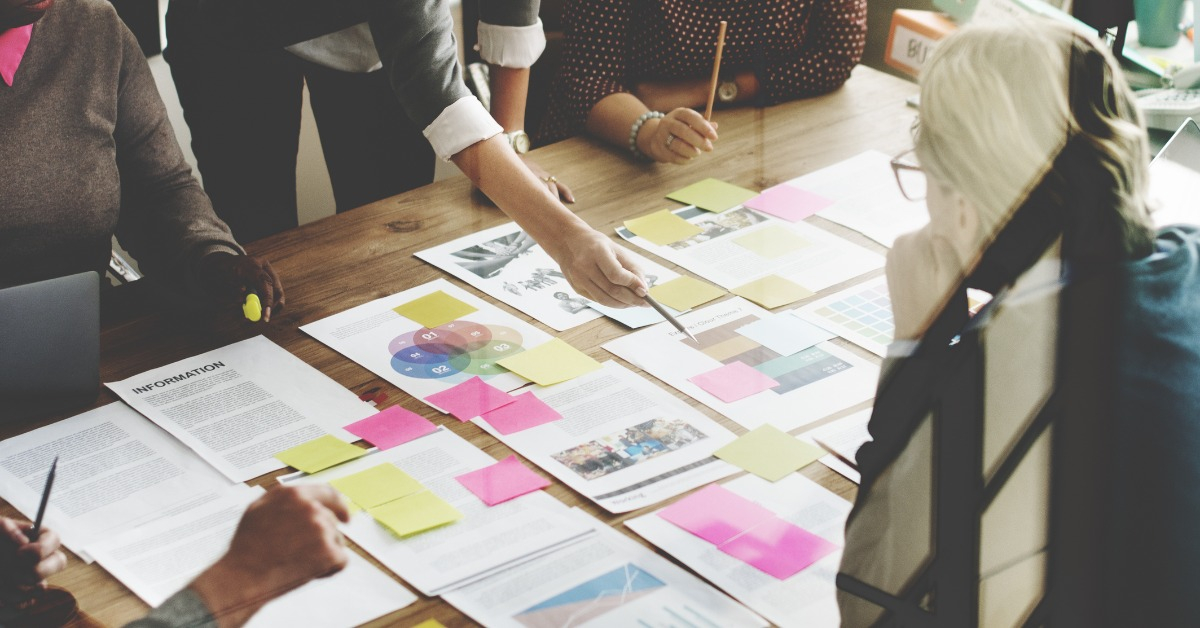 Team of people sitting around a desk with colorful non-descript papers laid out on the table with pink and yellow sticky notes attached to them. One person is pointing at a paper with a pencil in their hand. 