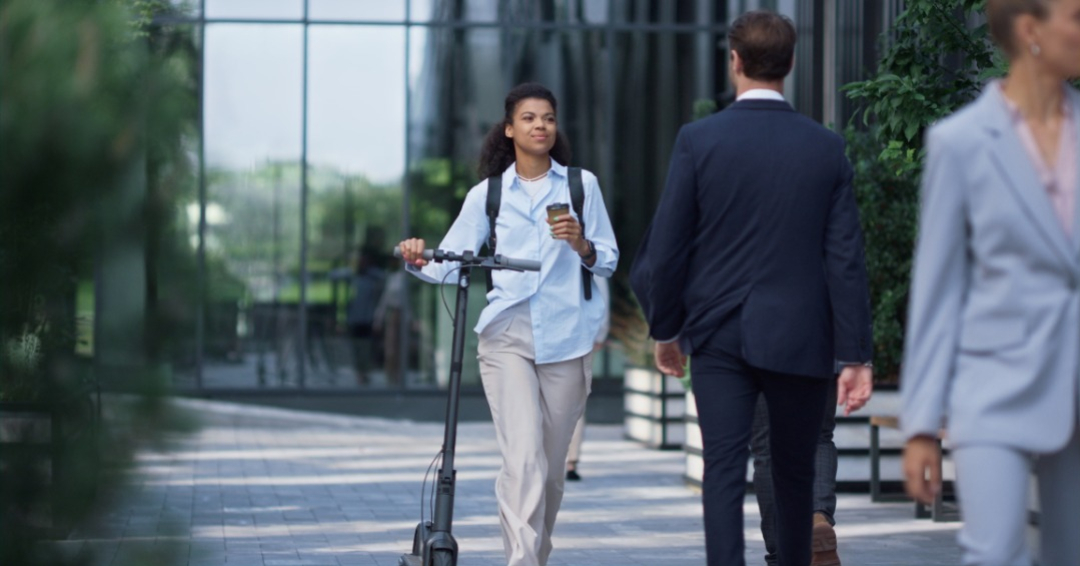 Smiling manager walking drinking coffee. Relaxed woman commuting home after work. Attractive african american woman worker going downtown holding electric scooter. Modern city transportation concept.