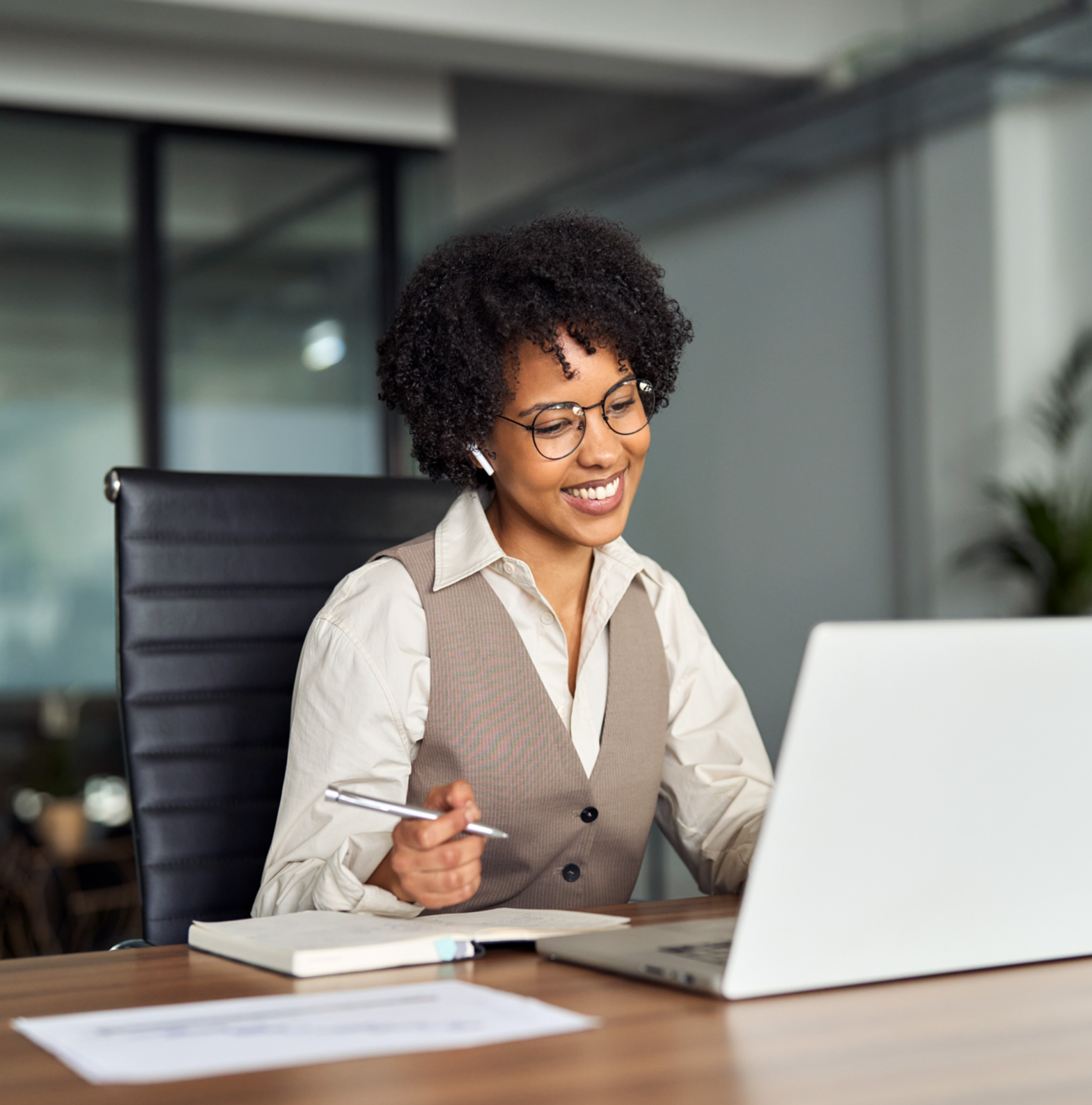 a woman in glasses with dark curly hair holds a pen and smiles at her open laptop