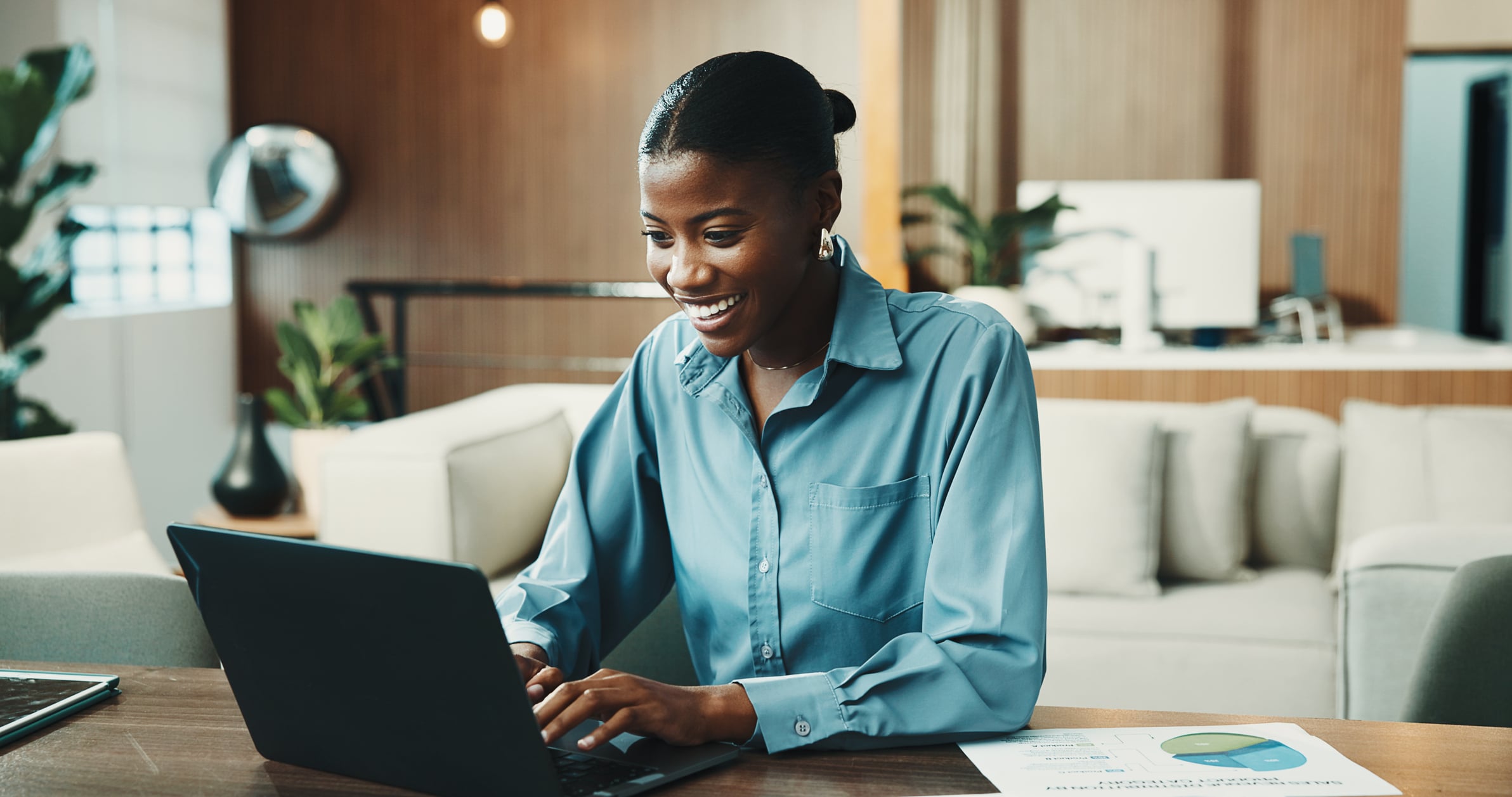 A professionally dressed woman smiles at her laptop screen with charts next to her