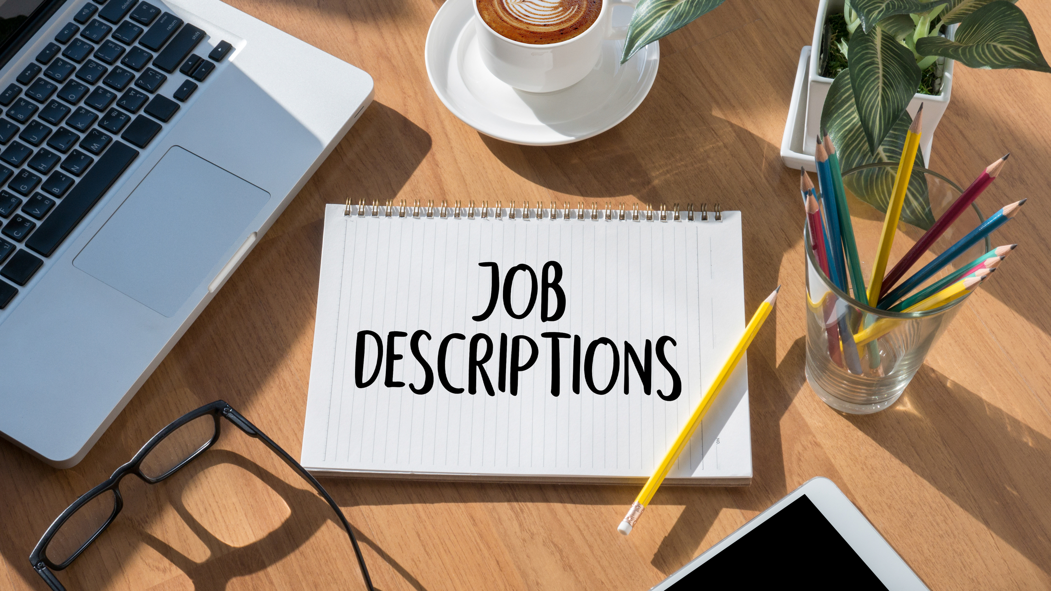 image of a notebook on a desk with the words Job Descriptions on it. On the desk is a cup of coffee on a saucer, a laptop, pencils in a cup, and glasses