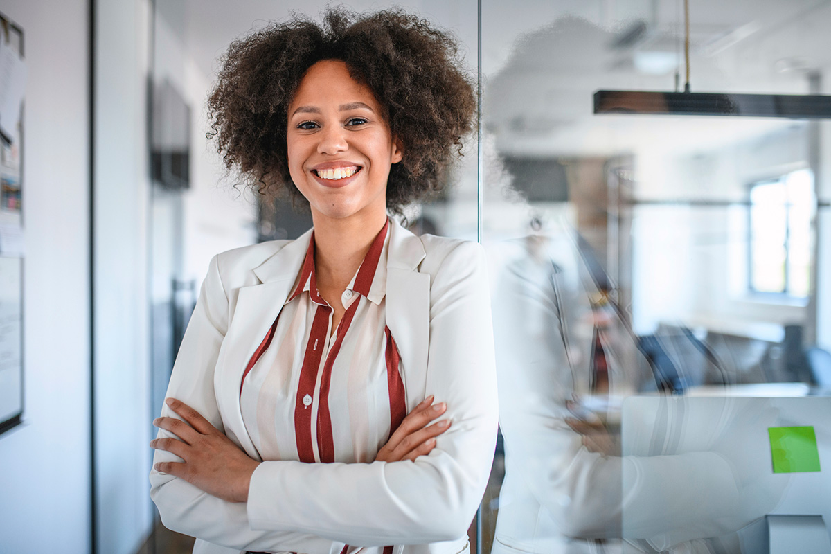 GettyImages-1162941402-Close-up of young smiling mixed race businesswoman standing confidently with arms crossed in a modern office.